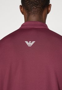 Bordo polo majica z teksturirano tkanino, z belim logotipom stiliziranih kril in inicialk na zadnjem ovratniku.