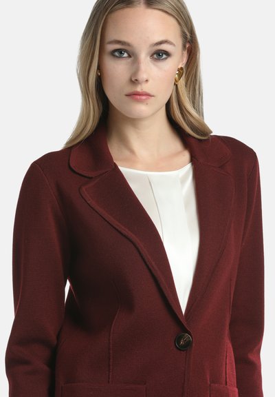 Woman with blonde hair wearing gold heart-shaped earrings, a white blouse, and a burgundy single-button blazer.