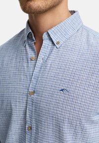 Camisa de cuadros azul claro y blanca, confeccionada en una tela de textura suave. Presenta un cuello, botones de madera y un detalle de logo bordado.