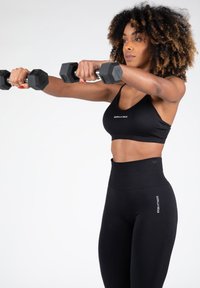 Sujetador deportivo negro y leggings de cintura alta fabricados con material elástico. La modelo sostiene mancuernas hexagonales, mostrando una silueta ajustada.
