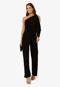 Combinaison noire à une épaule avec un détail noué à la taille, tissu fluide et coupe décontractée. Associée à des sandales à talons hauts.