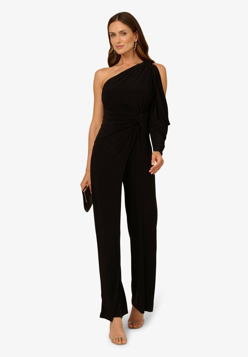 Combinaison noire à une épaule avec un détail noué à la taille, tissu fluide et coupe décontractée. Associée à des sandales à talons hauts.