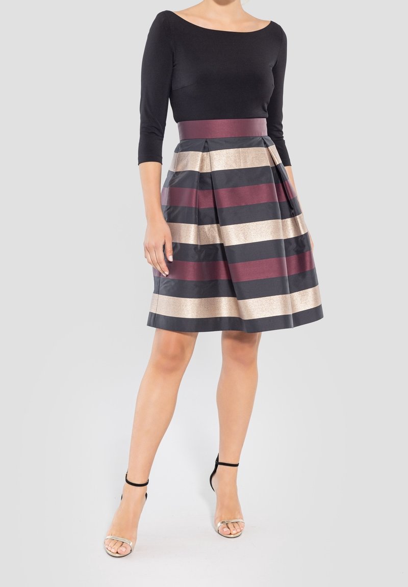 Femme portant un haut noir ajusté à manches trois-quarts et une jupe plissée à rayures horizontales noires, bordeaux et beige, ainsi que des sandales à talons hauts.