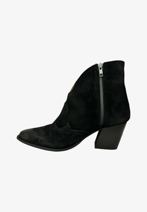 Botín de ante negro hasta el tobillo con punta afilada, detalles recortados y cremallera lateral, con un tacón grueso. Textura suave y diseño elegante.