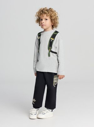 Bambino con capelli ricci in piedi che indossa una maglietta grigia a maniche lunghe con stampa di cinghie dello zaino, pantaloni neri con motivi grafici e sneakers bianche.