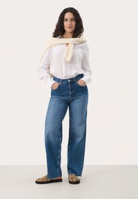 Jeans de mezclilla azul de pierna ancha combinados con una blusa blanca de manga larga. Un suéter crema está colgado sobre los hombros. Sandalias planas con detalles en marrón.