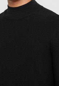 Pull noir côtelé à col haut porté sur le torse et le cou, montrant la texture tricotée et la coupe ajustée.