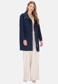 Marineblå yderjakke med lynlås foran, to brystlommer, trykknapper ved ærmerne og en struktureret hætte. Kombineret med lyse beige bukser med vidde.