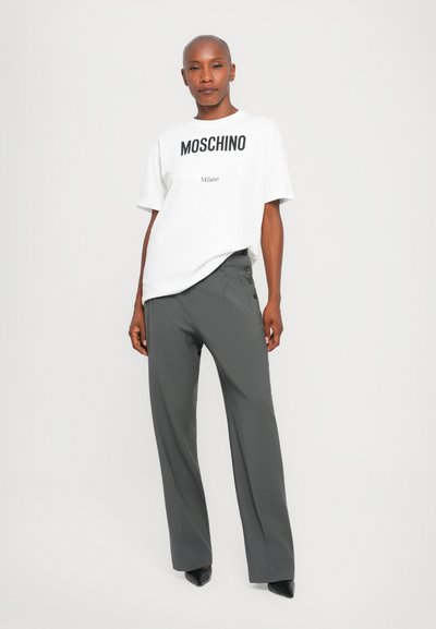Weißes Moschino T-Shirt mit schwarzem Logoschriftzug und grauen, weit geschnittenen Hosen mit Knopf-Details an der Seite, getragen mit spitzen schwarzen Schuhen.
