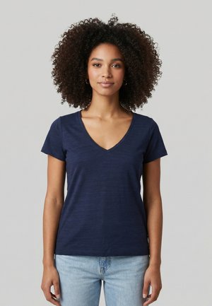 Donna con capelli ricci che indossa una T-shirt blu navy con scollo a V e jeans azzurri, in piedi davanti a uno sfondo grigio uniforme.