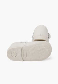Calzado blanco con un acabado liso y mate. Presenta una punta redondeada, un tacón bajo y una suela de goma con un patrón de agarre texturizado. Detalle de hebilla ajustable.