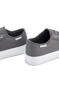 Gray canvas sneakers with a textured upper, white rubber sole, and side branding tag. Features round eyelets for laces.