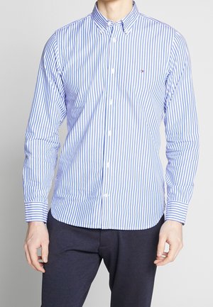 Hombre con camisa de manga larga a rayas verticales azules y blancas y pantalones oscuros, de pie contra un fondo liso.