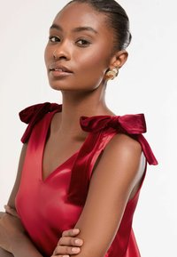 Vörös selyemruha, amelyet bársonyos masnik díszítenek a vállaknál, V kivágással. A sima textúra fokozza a elegáns dizájnt, a karok lazán összefonódva.
