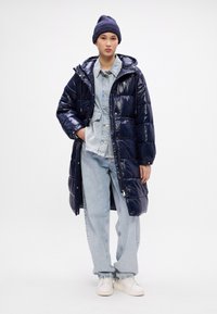 Palton puffer navy cu textură lucioasă, cu glugă și fermoare. Stratificat peste o jachetă ușoară din denim. Asortat cu blugi deschiși la culoare și adidași albi.
