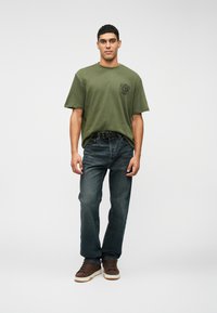 Camiseta de algodão verde-oliva com decote redondo e mangas curtas, combinada com jeans de ganga azul-escuro e ténis castanhos com soles brancos.