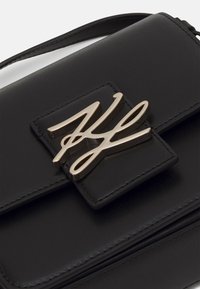 Czarna skórzana torebka z metalowym zapięciem w złotym odcieniu, prostokątnym kształcie, gładką fakturą oraz regulowanym paskiem na ramię.