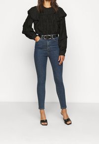 Blusa negra texturizada con mangas acampanadas, combinada con jeans azul de cintura alta. La modelo lleva sandalias negras tipo slide y un cinturón fino.
