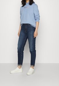 Lichtblauwe hoodie met koord en rood logo, gecombineerd met donkere spijkerbroek en witte sneakers, met een opgerolde manchette en slim fit.