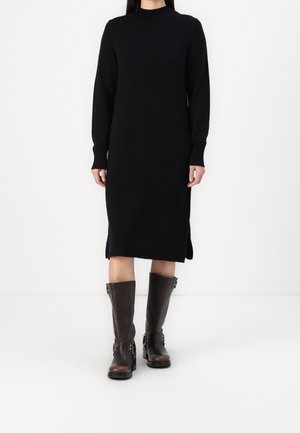 Vrouw draagt een zwart midi jurk met lange mouwen, gecombineerd met donkerbruine kniehoge leren laarzen met gespsdetails.