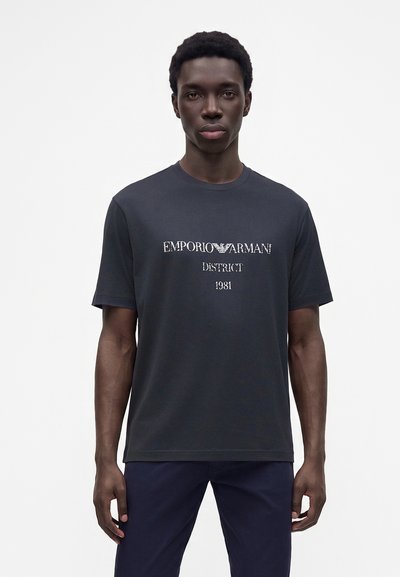 Νέος άνδρας που φοράει μαύρο κοντομάνικο T-shirt με εκτυπωμένο το "EMPORIO ARMANI DISTRICT 1981" στο μπροστινό μέρος, στέκεται μπροστά σε λευκό φόντο.