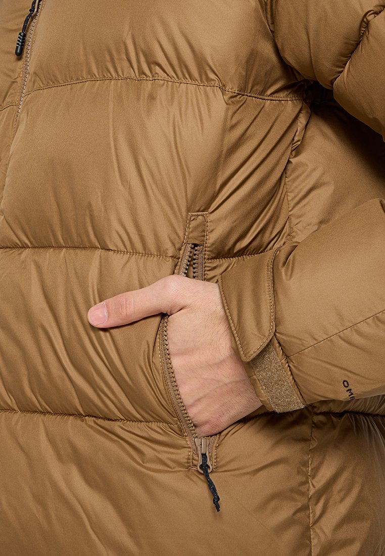 Main dans la poche latérale zippée d'une veste d'hiver matelassée marron avec une sangle en Velcro sur la manche.