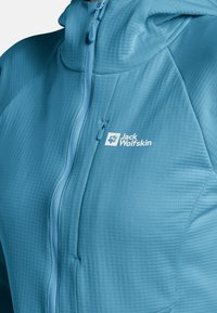 Veste Jack Wolfskin bleu clair avec une surface texturée, fermeture éclair intégrale, col montant et logo. Elle dispose d'un design ajusté et d'un tissu respirant.