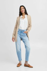 Béžový pletený cardigan přes bílou košili na knoflíky, světle modré džíny a hnědé mokasíny. Jednoduchý design s ribovanou texturou, volný střih.