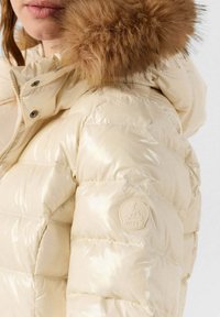 Chaqueta acolchada crema con acabado brillante, con capucha forrada de piel, textura acolchada y un logo circular en la manga.