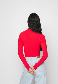 Rotes Langarmoberteil aus einem glatten, dehnbaren Stoff, kombiniert mit hellblauen Jeans, die auf der Rückseite ein sichtbares Markenetikett haben.