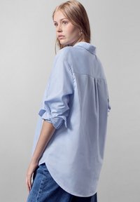 Camisa de manga larga azul claro con cuello y mangas enrolladas. Presenta un dobladillo redondeado y un corte relajado con un detalle de costura en el yugo trasero.