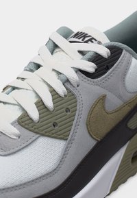 Primo piano di una sneaker Nike grigia, bianca e oliva con lacci bianchi e materiali in tessuto strutturato e pelle scamosciata.