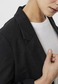 Černý blazer s texturovanou tkaninou, s klopami a viditelným bílým tričkem pod ním. Upravuje límec rukou.