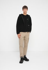 Un sweat à capuche en polaire noire avec une fermeture éclair à l'avant, associé à un t-shirt noir et un pantalon cargo beige. Des chaussures noires imposantes complètent la tenue.