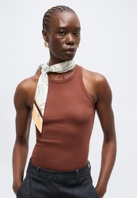 Femme aux cheveux tressés, portant un haut sans manches couleur rouille, un pantalon noir, des boucles d’oreilles dorées et un foulard blanc à motifs autour du cou.