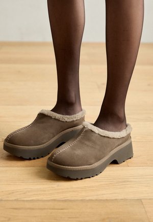 Pieds portant des sabots marron à enfiler avec une semelle épaisse et une garniture en fourrure claire, associés à des bas noirs transparents sur un sol en bois.