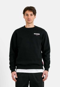 Zwarte sweatshirt met lange mouwen, geribbelde manchetten en zoom. Heeft een klein logo op de borst en is over een wit shirt gedragen.