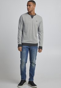 Grauer Pullover mit schwarzem Kragen und Bündchen, kombiniert mit blauen Jeans. Das Modell trägt schwarze Sneakers und steht vor einem schlichten Hintergrund.