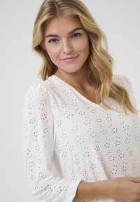 Blouse blanche avec un design en œillets, encolure ronde et manches longues bouffantes. Présente des motifs brodés et une texture légère et respirante.