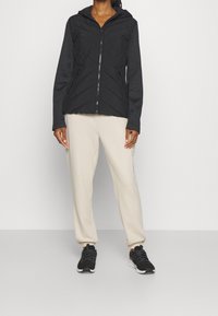 Czarna kurtka na zamek z kapturem, ozdobiona pikowanymi wzorami w kształcie jodełki. W zestawie z jasnobeżowymi spodniami i czarnymi butami sportowymi.