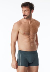 Boxers ajustados de color verde oscuro, hechos de una tela suave, con un ajuste ceñido, cinturilla elástica y ribetes laterales blancos. Sin patrones ni logotipos visibles.