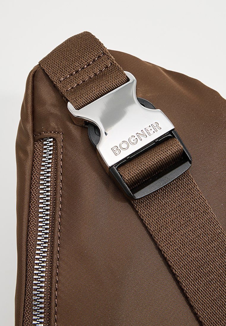 Brauner Stofftaschenriemen mit silbernem Metallschnallen, graviert mit "BOGNER", und einem sichtbaren Reißverschluss auf der linken Seite.