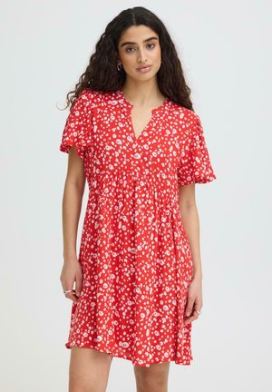 Mladá žena s dlhými kučeravými vlasmi, oblečená v krátky červený šaty s bielym kvetinovým vzorom a rozšírenými rukávmi, stojaca na jednoduchom pozadí.