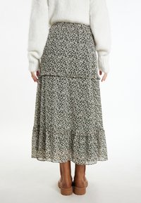 Florale maxi-rok met een groene basis, voorzien van witte bloempatronen, gelaagde ruffles en een elastische tailleband. Bruine kniehoge laarzen worden gedragen.