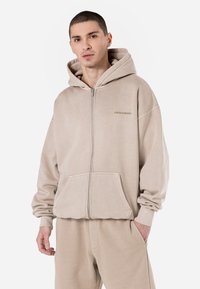Beige zip-up hoodie lavet af blødt stof, med en afslappet pasform, kængurulommer og mærket "PGA" broderet på brystet.