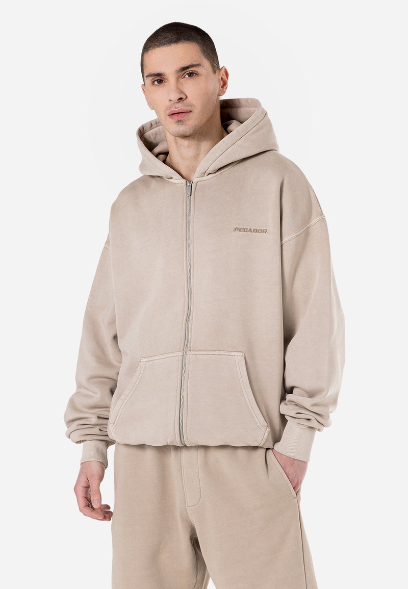 Felpa beige con zip realizzata in tessuto morbido, caratterizzata da una vestibilità comoda, tasche a marsupio e il nome del marchio "PGA" ricamato sul petto.
