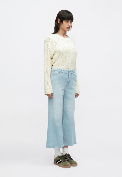 Πλεκτό πουλόβερ σε ανοιχτό μπεζ με υφή, συνδυασμένο με φαρδιά ανοιχτό μπλε τζιν παντελόνια και πράσινα αθλητικά παπούτσια με μαύρες ρίγες.