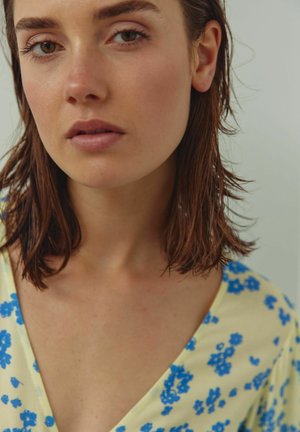 Portrét osoby so stredne dlhými hnedými vlasmi, ktorá má na sebe žltý vrchný odev s kvetinovým vzorom modrých kvetov a hlbokým výstrihom do V.
