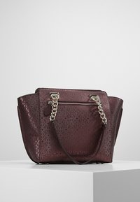 Un bolso burdeos con una superficie texturizada, que presenta un asa de cadena y cremallera, se encuentra sobre un pedestal gris claro contra un fondo liso.