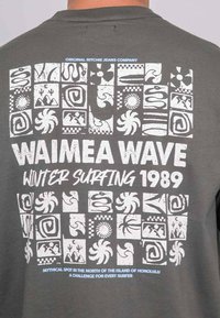 T-shirt gris avec impression graphique blanche présentant des motifs abstraits et le texte "WAIMEA WAVE WINTER SURFING 1989" au dos.